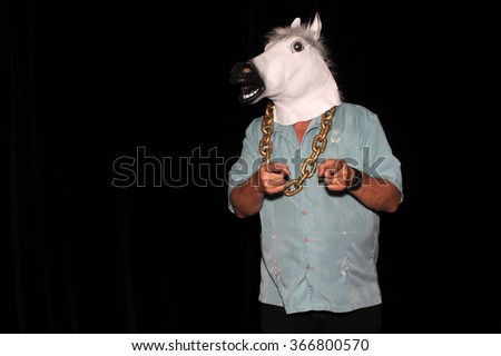 An unidentifiable man wears a White Rubber Horse Head Mask while wearing a Gold Chain and points to the camera in a Photo Booth. With Black velvet curtains and room for your text