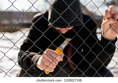 Unidentifiable Teenage Boy Behind Wired Fence Holding A Paper Knife At Correctional Institute, Focus On The Fence, Conceptual Image Of Juvenile Delinquency.