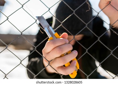 Unidentifiable Teenage Boy Behind Wired Fence Holding A Paper Knife At Correctional Institute, Focus On The Boys Hand, Conceptual Image Of Juvenile Delinquency.