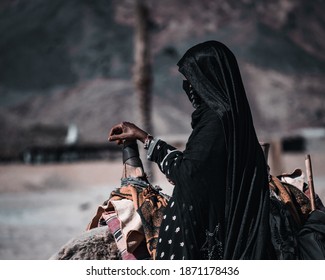 An Unidentified Bedouin Woman In A Black Abaya Standing Near A Lying Camel With Her Hand On A Traditional Camel Saddle