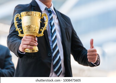 Unidentified Business People Holding The Golden Trophy Cup For Their Success Winner In Business Project.  The Best Business Man Has Achieved Their Desired Gold With Golden Trophy In Middle City Town.