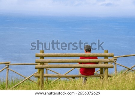 unidentified man with his back turned, wearing a red shirt, is sitting on a wooden bench, watching the Atlantic Ocean on the island of Santa Maria, Azores, Portugal.
