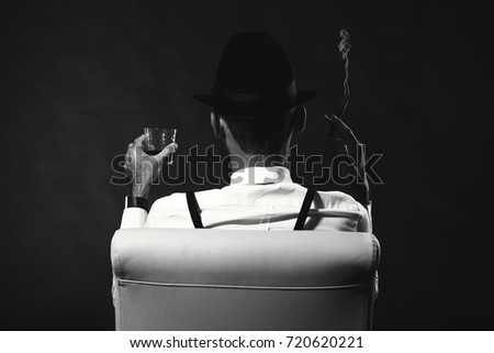 an unidentified man in a suit and hat with a cigar and whiskey staring into the distance. View from the back. The man is a mafia. Millionaire gangster businessman boss