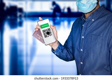 unidentified man wearing a face mask and holding a passport and a Green pass with meaningless QR code representing a certificate of vaccination.Airport on the background.