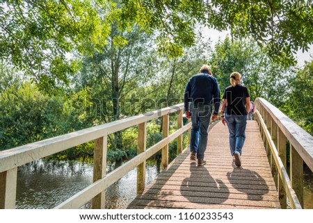 An unidentified  man and woman in casual clothes and shoes walk over a simple wooden bridge over a narrow river on a sunny day in the Dutch summer season.