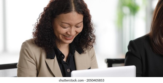 Unidentified Unrecognizable Asian Middle Aged Female Businesswoman In Formal Suit Sitting Working Typing Notebook Laptop Computer Together Side By Side On Table Full Of Report Paperwork Document.