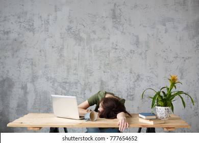 Unidentified young girl office worker fell asleep while working at desk with laptop overturned mug on background of gray vintage wall. Place for text or ads