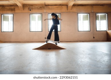 Unidentified young woman in casual sports clothing learning skateboarding trying to balance with arms extended on artificial indoor small slide in skating studio during daytime