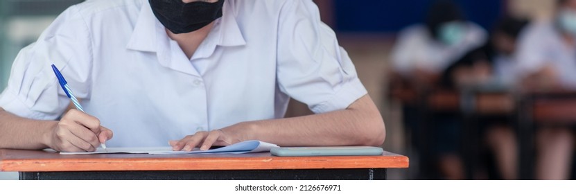 Uniform Student Taking Exam While Wearing Face Mask Due To Coronavirus Emergency. Young Woman Sitting In Class With Wearing Surgical Mask Due To Covid-19 Pandemic