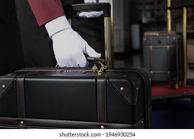 A Uniformed Doorman Holds A Brown Leather Suitcase In His Hand. The Hands Of The Doorman In White Cotton Gloves.Service At The Hotel .The Holiday Season.