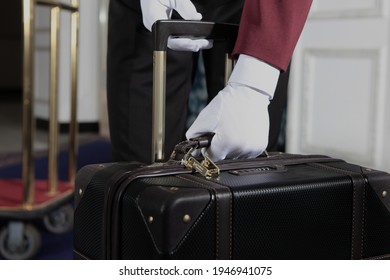 A Uniformed Doorman Holds A Brown Leather Suitcase In His Hand. The Hands Of The Doorman In White Cotton Gloves.Hotel Service .The Holiday Season.An Unrecognizable Person.Luggage Trolley At The Hotel.