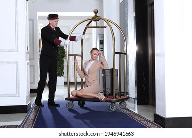 A Uniformed Doorman Pushes A Luggage Cart Toward The Elevator.A Young Woman Sits On A Luggage Cart.Hotel Service.The Holiday Season. Business Woman On A Business Trip.