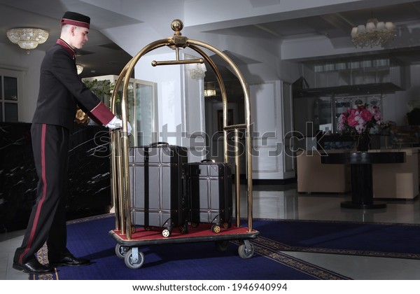 A uniformed doorman pushes a luggage cart .Several\
leather suitcases stand on a luggage cart. Hotel service.The\
holiday season. Free space.