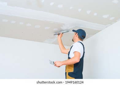 A uniformed worker applies putty to the drywall ceiling. Putty of joints of drywall sheets. - Shutterstock ID 1949160124