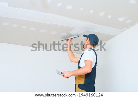 A uniformed worker applies putty to the drywall ceiling. Putty of joints of drywall sheets.