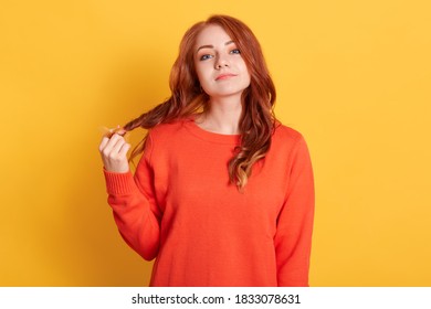 Unimpressed Indifferent Attractive Woman With Red Hair, Pulling Her Curls, Feeling Bored, Being On Boring Date, Standing Annoyed And Tired Over Yellow Background, Wearing Orange Jumper.