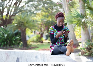 Uninhibited Young African Teenager In Modern Clothes And Dyed Frizzy Hair Siting Barefoot With Legs Outstretched In The Park Exchanging Messages With Her Friend On Her Smartphone