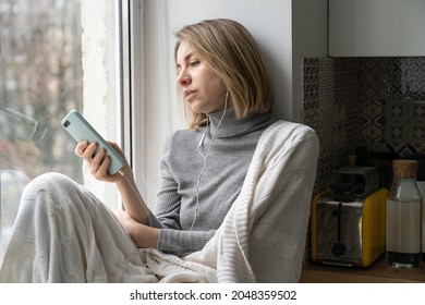 Uninterested woman wrapped in plaid sitting on windowsill at home, not in mood, using smartphone, wear wired headphones and listens music or podcast. Bored female addicted and chatting in social media - Powered by Shutterstock