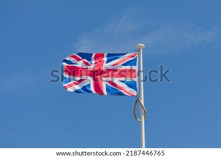 Union Jack flag on flagpole in strong breeze, flying to the left, left side is facing camera, blue sky with small cloud.