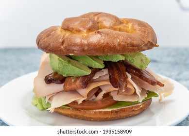 Unique Chicken Club Sandwich Served On A Pretzel Bun And Loaded With Avocado Wedges, Bacon, Meat, And Lettuce.