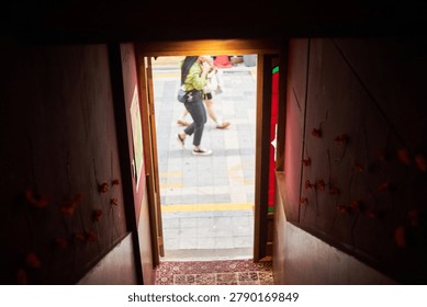          A unique and cinematic perspective looking from inside a dimly lit, atmospheric cafe or bar out onto a bright city street. The narrow entrance features dark red walls decorated with delicate floral ornaments, leading to a wooden doorway with a patterned rug on the floor. Through the door frame, anonymous pedestrians are captured walking past the entrance. In one scene, a man in a business suit walks by, while in another, casually dressed individuals stroll along the pavement. The intentional blurring of the people outside contrasts sharply with the focused, dark interior, creating a voyeuristic and moody storytelling image that perfectly captures the fleeting moments of everyday urban life.                      