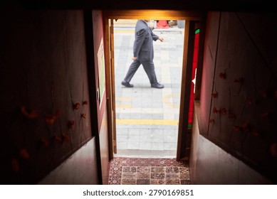          A unique and cinematic perspective looking from inside a dimly lit, atmospheric cafe or bar out onto a bright city street. The narrow entrance features dark red walls decorated with delicate floral ornaments, leading to a wooden doorway with a patterned rug on the floor. Through the door frame, anonymous pedestrians are captured walking past the entrance. In one scene, a man in a business suit walks by, while in another, casually dressed individuals stroll along the pavement. The intentional blurring of the people outside contrasts sharply with the focused, dark interior, creating a voyeuristic and moody storytelling image that perfectly captures the fleeting moments of everyday urban life.                      