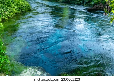 Unique karst source of water gushing from the ground called the Blue Eye or Syri i Kalter. Natural phenomenon occurring in the village of Muzine, Albania. River framed by green trees.