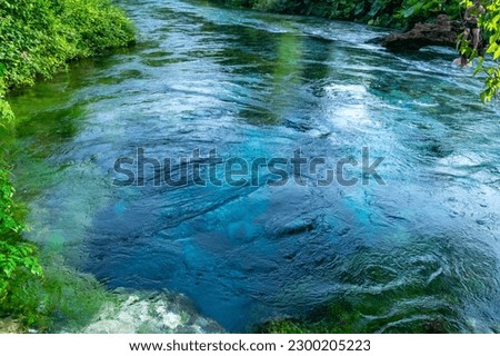 Unique karst source of water gushing from the ground called the Blue Eye or Syri i Kalter. Natural phenomenon occurring in the village of Muzine, Albania. River framed by green trees.