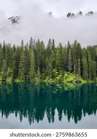 A unique mountain lake reflection at Lake Carezza in the Dolomites, Italy, with a dense layer of fog drifting above dark fir trees. The mirror-like water surface captures the mysterious atmosphere.
