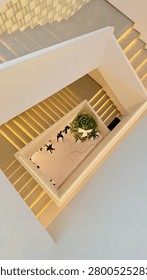 Unique overhead perspective of a modern multi-level staircase featuring clean lines, warm ambient lighting, and decorative indoor plants. Contemporary architectural interior with geometric shapes and minimalist aesthetics, ideal for themes related to luxury buildings, interior design, and modern living spaces.