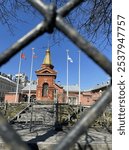 A unique perspective of a historic building with a golden spire, framed by a chain-link fence. Flags fly against a clear blue sky, adding a patriotic touch to the architectural scene