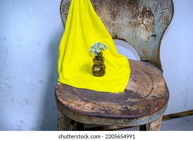 A unique rustic flare using a yellow wash cloth and an antique metal chair. The display is set in front of a weathtered and worn wall. Slight defocus effect points attention to the flower.