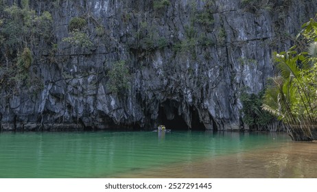 A unique underground river. A canoe with tourists at the entrance to a dark cave. Sheer cliffs rise above the calm emerald water.  Palm trees by the shore. Green vegetation on steep slopes.Philippines - Powered by Shutterstock