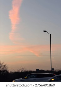 Unique vertical pink cloud formation in the twilight sky above a road with a street lamp silhouette. Minimalist urban evening scene featuring pastel colors and soft sunset light.