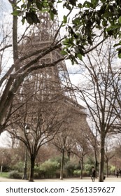A unique view of the Eiffel Tower in Paris, France, framed by bare winter trees under a bright, partly cloudy sky. The image captures the iconic iron structure from the ground while people enjoy the