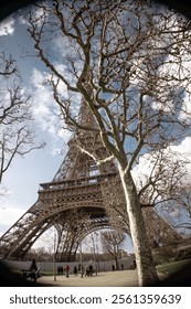 A unique view of the Eiffel Tower in Paris, France, framed by bare winter trees under a bright, partly cloudy sky. The image captures the iconic iron structure from the ground while people enjoy the