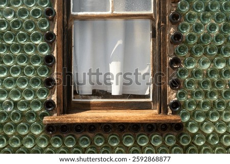Unique wall made of embedded green and brown glass bottles surrounding a rustic wooden window with white curtains. Ideal for architecture, sustainability, and creative design themes.