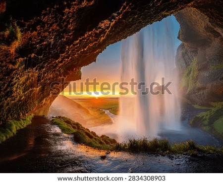 Unique wide-angle interior perspective looking out from behind the cascading curtain of Seljalandsfoss waterfall in South Iceland. Vibrant golden sunset rays filter through the misty water spray, illuminating the mossy green cavern walls and surrounding coastal plains under an amber sky. Perfect for Icelandic Golden Circle tours, nature adventure promos, and high-impact landscape photography.