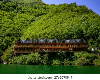 Unique wooden hotel structure with angular roofs integrated into a vibrant forested mountain slope, overlooking the shimmering green water of Shala River, Koman Lake, Albania. Bright summer daylight.