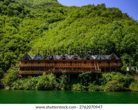 Unique wooden hotel structure with angular roofs integrated into a vibrant forested mountain slope, overlooking the shimmering green water of Shala River, Koman Lake, Albania. Bright summer daylight.
