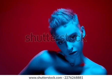 Uniqueness. Beautiful young girl with short blonde hair posing with bare shoulders against red studio background in blue neon light. Cyberpunk style. Concept of futurism, digital world, robot, art