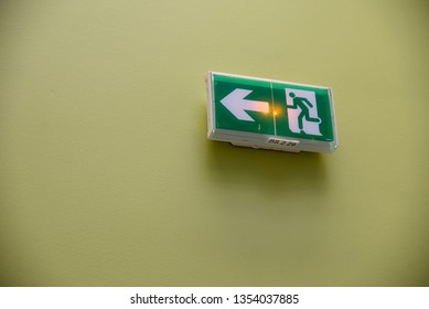 Universal Exit Signage: An Exit Sign With A Directional Arrow And Internal Emergency Lighting On A Plain Green Painted Wall