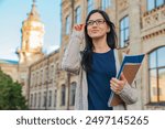 University college female student woman in eyeglasses casual clothes smiling with with books notebooks and bag standing outside near on campus building outdoor and looking away. Education concept