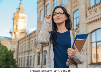 University college female student woman in eyeglasses casual clothes smiling with with books notebooks and bag standing outside near on campus building outdoor and looking away. Education concept