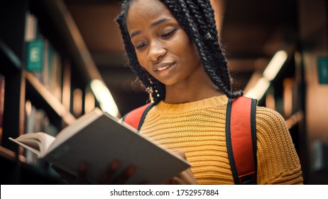 University Library: Smart Beautiful Black Girl Standing Next to Bookshelf Holding and Reading Text Book, Doing Research for Her Class Assignment and Exam Preparations. Low Angle Portrait with a Smile
