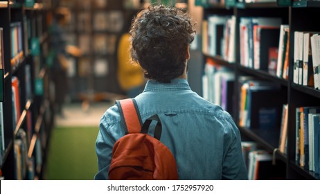 University Library: Student Walks Between Rows Of Bookshelves Searching For The Right Book Title For Class Assignment And Exam Preparations. Back View Shot. Young People Study, Learn