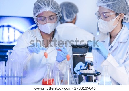 University science students learning practical chemistry experiment. Young researchers in protective uniforms studying red liquid reaction in laboratory room site. Professional biotechnology experts analyzing DNA structure on digital screens. Scientists in safety gear conducting microscopic research for medical innovation. Collaborative medical research team working together in modern science laboratory. Male and female scientists analyzing red chemical sample with microscope tool.