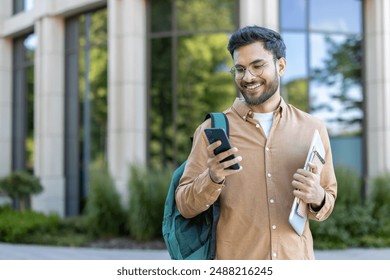 University student standing outside modern building smiling while looking at phone. Student carrying backpack and folder, representing education, technology, and youth lifestyle on campus. - Powered by Shutterstock