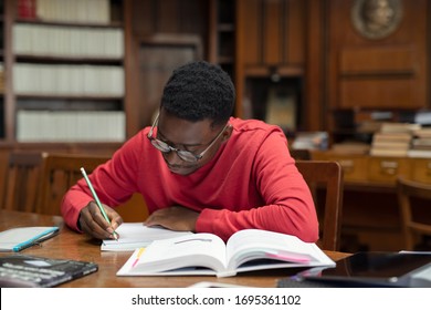 University Student Wearing Spectacles Studying In Library. Young African Man Taking Notes From Book While Sitting In The Library. Focused Casual Guy Writing Notes During Homework.