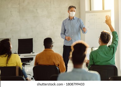 University Teacher With Protective Face Mask Pointing At Student With Raised Arm During A Class At Lecture Hall. 